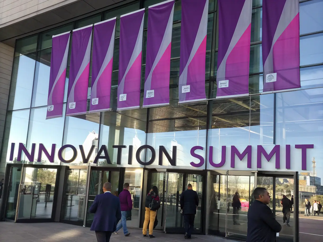 A photograph of the entrance to a modern conference center in London, with banners advertising an 'Innovation Summit'. The image should convey excitement and anticipation.